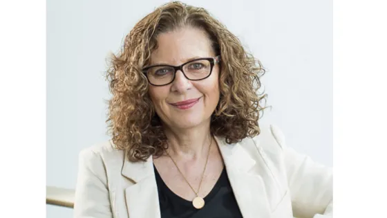 A woman with curly hair and glasses, smiling while seated in a white blazer.