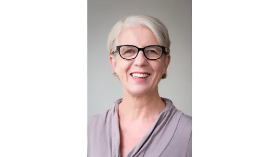 A portrait of an elderly woman with short white hair, smiling, wearing glasses and a lavender blouse, against a light gray background.
