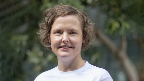 A woman smiling at the camera with short wavy hair, wearing a white t-shirt, with a blurred green background.