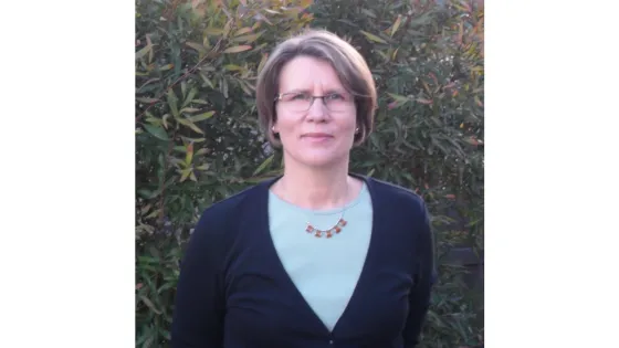 A woman wearing glasses, a green shirt, and a black cardigan stands in front of a leafy bush.