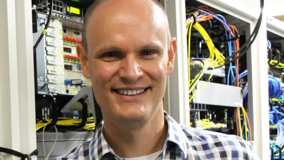 A smiling man in a plaid shirt standing in front of server racks in a data center.