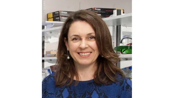 A woman with brown hair, smiling, wearing a blue blouse, standing in a laboratory environment with shelves in the background.