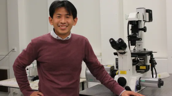 A man smiling at the camera stands beside a microscope in a laboratory setting.