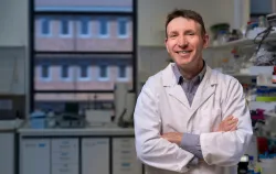 A smiling man in a lab coat standing with crossed arms in a laboratory.