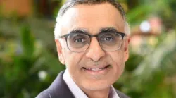 A portrait of a smiling middle-aged man wearing glasses, with graying hair, dressed in a professional blazer against a blurred green background.