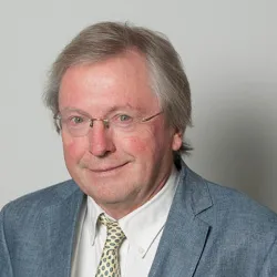 A portrait of an elderly man with glasses, smiling, wearing a light blue blazer and a patterned tie against a light gray background.