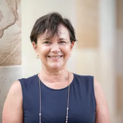 A smiling middle-aged woman wearing a black sleeveless dress and a necklace stands in front of a textured wall.