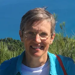 A smiling woman with short hair, wearing glasses, a blue jacket, and a red shoulder bag, stands outdoors with a blue sky and green vegetation in the background.
