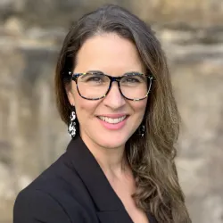 A smiling woman wearing glasses and patterned earrings stands before a blurry stone wall background.