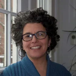 A smiling woman with curly gray hair wearing glasses and a blue scarf stands near a window.