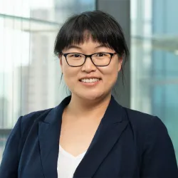 A portrait of a smiling woman with glasses wearing a dark blue blazer, standing in front of a glass-paneled office background.
