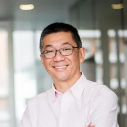 A man in a pink shirt smiling at the camera in a bright office hallway.