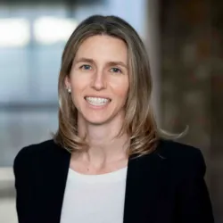 A professional portrait of a smiling woman with long blond hair, wearing a black blazer and white top, standing in a modern office setting.