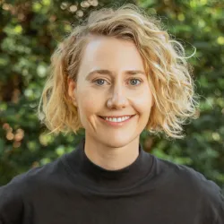 A portrait of a smiling woman with short curly blonde hair, wearing a black turtleneck, standing before a leafy green background.