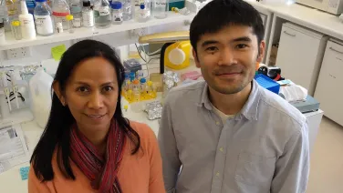 Two scientists, a man and a woman, smiling in a laboratory filled with scientific equipment and bottles.