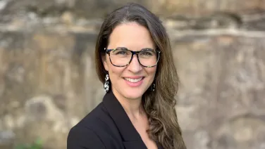 A smiling woman wearing glasses and patterned earrings stands before a blurry stone wall background.